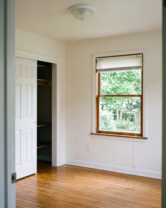 An empty bedroom with white walls, honey oak floors, an open closet and a double-hung window looking out at green trees