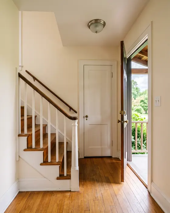 An empty entry hall with a wooden staircase and white banister, honey oak floors, a closet door and the front door open to a porch and green trees