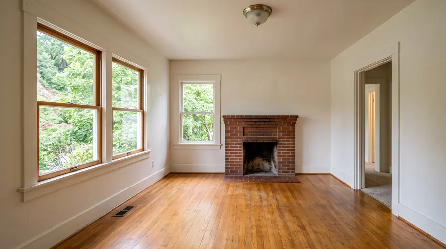 An empty living room with honey oak floors, a brick fireplace and tall wood-framed windows looking out at green trees